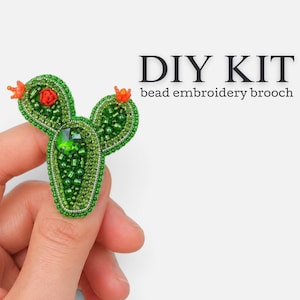 Può includere: Un kit fai-da-te per una spilla ricamata con perline a forma di cactus, con perline di vetro verde, un cristallo verde sfaccettato centrale e piccoli accenti floreali arancioni. Il testo DIY KIT bead embroidery brooch è stampato in nero a destra.