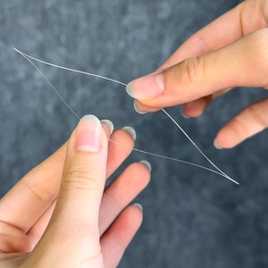 May include: Close-up of two hands holding and manipulating thin, white threads against a gray background. The threads are arranged in a geometric shape, possibly for crafting or repair purposes. The image highlights the delicate nature of the material.