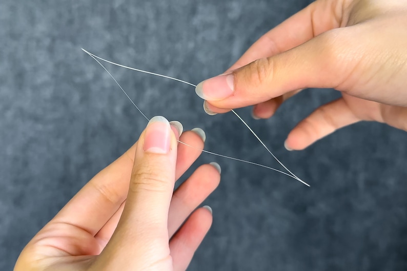 May include: Close-up of two hands holding and manipulating thin, white threads against a gray background. The threads are arranged in a geometric shape, possibly for crafting or repair purposes. The image highlights the delicate nature of the material.