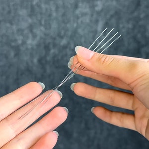 May include: Close-up of several thin, silver-colored wires held between two hands. The wires are arranged in a fan-like pattern, with some strands separated and others bundled together. The background is a dark, textured surface.