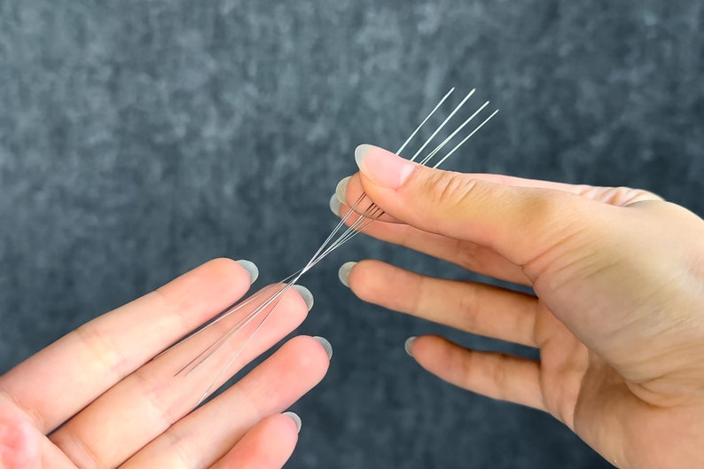 May include: Close-up of several thin, silver-colored wires held between two hands. The wires are arranged in a fan-like pattern, with some strands separated and others bundled together. The background is a dark, textured surface.