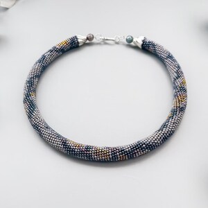 May include: A beaded necklace with a tubular design. The necklace features a pattern of small, multicolored beads in shades of gray, black, and gold. The necklace is approximately 16 inches in length and has a silver clasp.