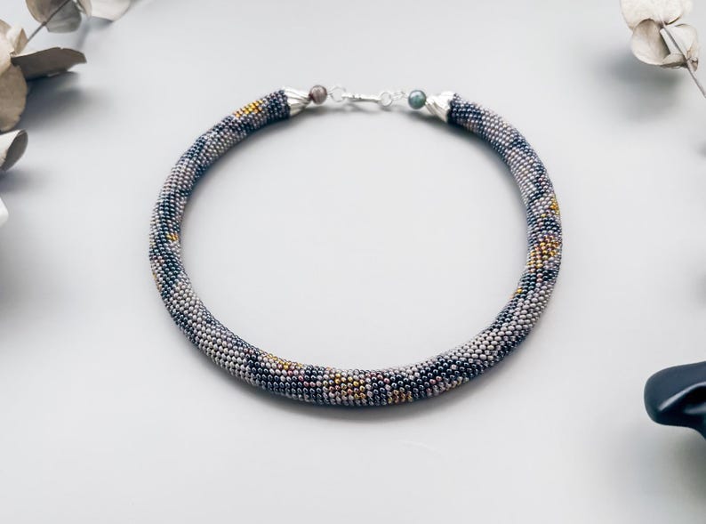 May include: A beaded necklace with a tubular design. The necklace features a pattern of small, multicolored beads in shades of gray, black, and gold. The necklace is approximately 16 inches in length and has a silver clasp.