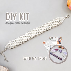Może przedstawiać: Zestaw DIY do wykonania bransoletki ze smoczej łuski, zawierający gotową białą bransoletkę z koralików ze srebrnym zapięciem typu karabińczyk i łańcuszkiem przedłużającym. Zestaw zawiera drukowaną instrukcję oraz okrągłą wstawkę z koralikami.
