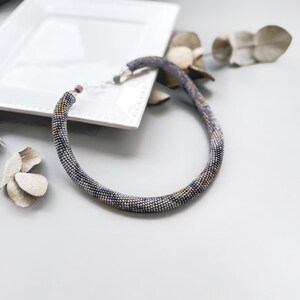 May include: A beaded necklace in shades of gray, blue, and gold, resting on a white square plate. The necklace is a choker style, with a beaded design. Dried leaves and a white plate are in the background.