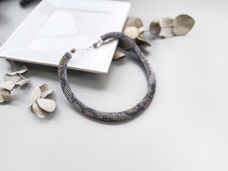 May include: A beaded necklace in shades of gray, blue, and gold, resting on a white square plate. The necklace is a choker style, with a beaded design. Dried leaves and a white plate are in the background.