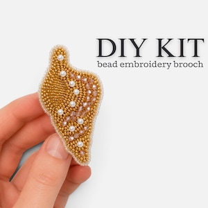 以下が含まれることがあります： 貝殻の形をしたビーズ刺繍ブローチのDIYキット。このブローチは、ゴールドトーンのシードビーズ、小さな白いパール、ライトブラウンのファセットカットクリスタルをテクスチャーのあるパターンで配置して作られています。DIY KIT bead embroidery broochというテキストが表示されています。