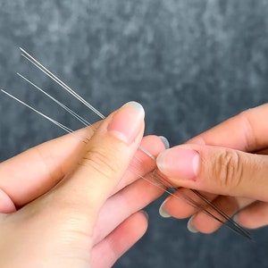 May include: Close-up of a person's hands holding several thin, white, and translucent strands. The strands are arranged in a fan-like pattern, with some strands appearing to be slightly longer than others. The background is a solid, muted gray.