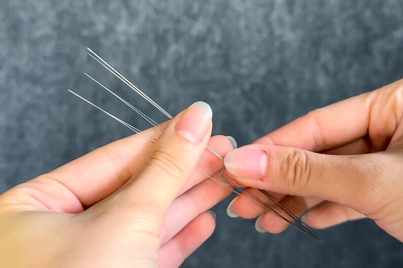 May include: Close-up of a person's hands holding several thin, white, and translucent strands. The strands are arranged in a fan-like pattern, with some strands appearing to be slightly longer than others. The background is a solid, muted gray.