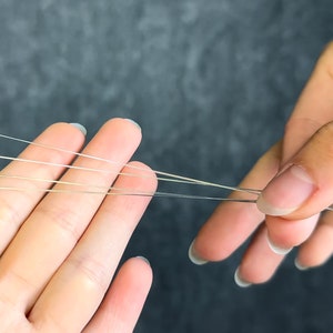 May include: Close-up of several thin, silver-colored needles held between two hands. The needles are straight and have sharp points. The background is a solid, dark gray color.