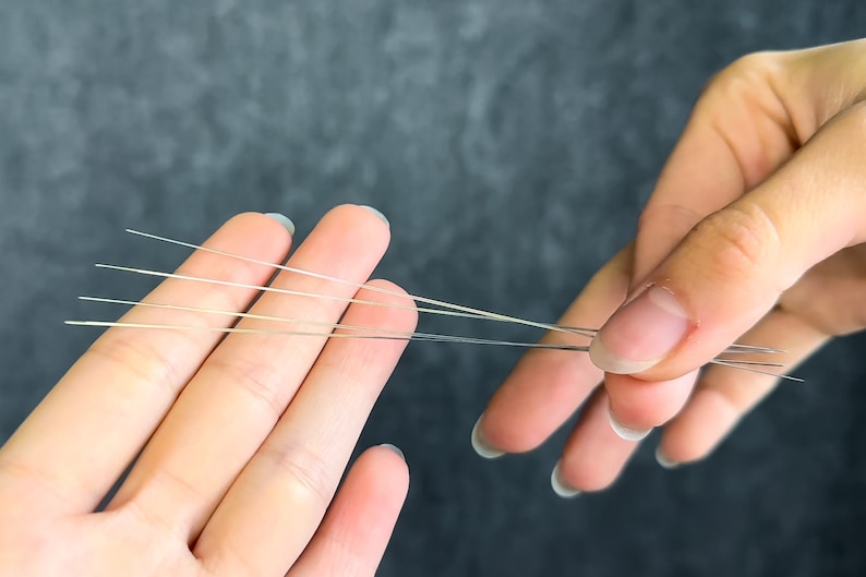 May include: Close-up of several thin, silver-colored needles held between two hands. The needles are straight and have sharp points. The background is a solid, dark gray color.