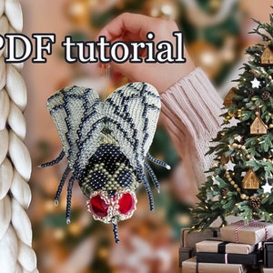 May include: A beaded fly ornament with black, white, and silver beads, and red eyes. The ornament is being held by a hand in front of a Christmas tree decorated with wooden houses and stars.