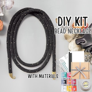 May include: A DIY bead necklace kit featuring a long, black tubular beaded rope with gold-coloured end caps. The kit includes a printed pattern guide, a small cardboard gift box with a grey ribbon, various small bags of colourful beads, and a spool of thread.