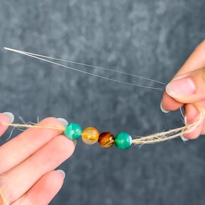 May include: A strand of colorful gemstone beads in shades of green, orange, and brown, strung on a natural twine cord. The beads are being strung with a needle and thread, suggesting a DIY jewelry project.