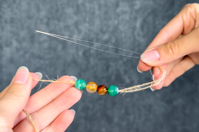 May include: A strand of colorful gemstone beads in shades of green, orange, and brown, strung on a natural twine cord. The beads are being strung with a needle and thread, suggesting a DIY jewelry project.