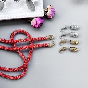 Peut inclure: Un collier de perles rouges avec des embouts en m&eacute;tal dor&eacute; et argent&eacute;. Le collier est dispos&eacute; en forme de c&oelig;ur sur une surface grise.