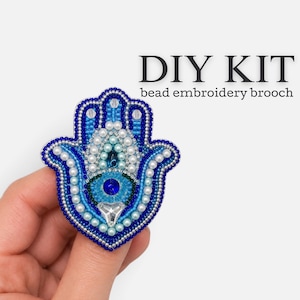 May include: A DIY kit for a bead embroidery brooch in the shape of a Hamsa hand. The brooch features intricate blue, light blue, and white beads, with a central dark blue stone and a clear triangular crystal accent at the bottom base.