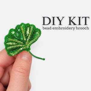 May include: A ginkgo leaf-shaped bead embroidery brooch DIY kit featuring various shades of green beads, including small round beads and elongated crystals, arranged in a fan-like pattern with a thin beaded stem and the text DIY KIT bead embroidery brooch.