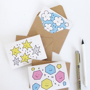 May include: Two greeting cards with hand-drawn illustrations. The first card features yellow stars with smiling faces and the text "en, P, 0, ph, Ins". The second card features colorful geometric shapes with smiling faces and the text "d, P, 0, a, m, i, n, e".