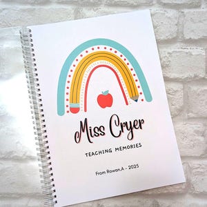May include: A white spiral-bound notebook with a rainbow illustration, a red apple, and the text "Miss Cryer TEACHING MEMORIES". The notebook is personalized with the name "From Rowan.A - 2025". The cover is against a white brick wall.