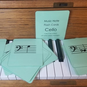 Puede incluir: Tarjetas de notas musicales verde claro para violonchelo, con notación musical negra, colocadas en un teclado de piano. Las tarjetas muestran el texto "Music Note Flash Cards Cello" y "notes in first position with letter names and fingers."