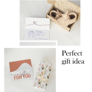 May include: A gift set featuring baby shoes in a box, a card with handwritten text, and a gift card. The shoes are light pink. The gift card says "A Gift For You". The wrapped gift has a floral pattern.