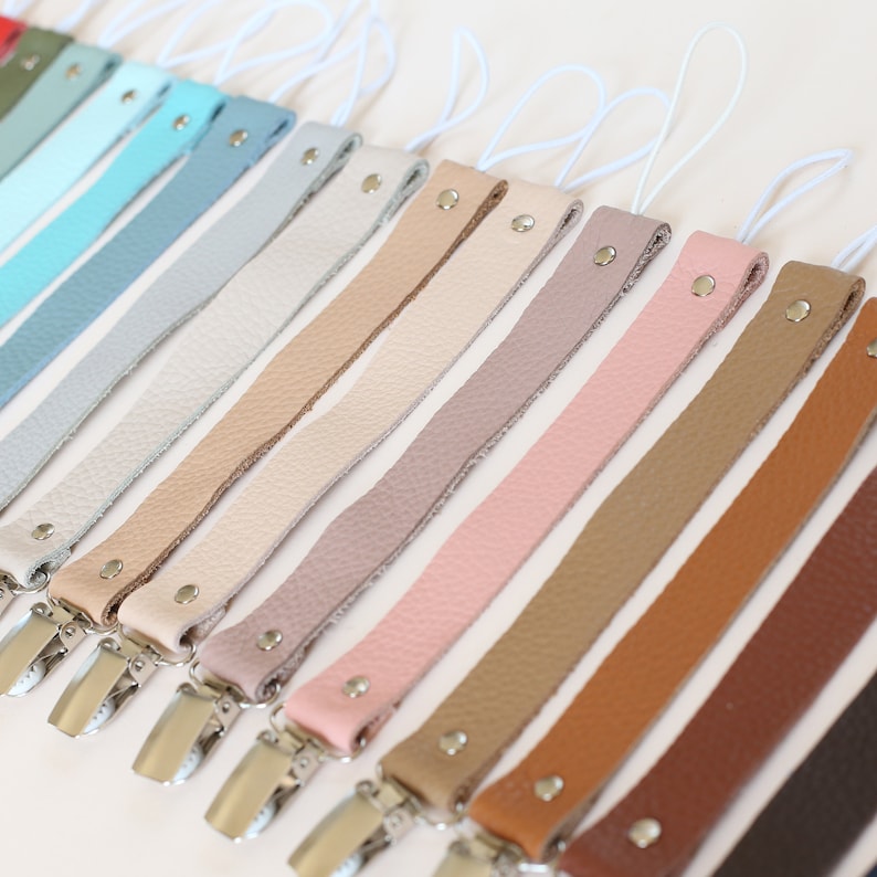 Puede incluir: Una colecci&oacute;n de 12 clips para chupetes de cuero en varios colores, incluyendo azul claro, beige, marr&oacute;n y rosa. Cada clip tiene un cierre de metal plateado.