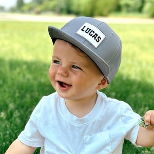 May include: A smiling baby wearing a grey baseball cap with a white patch that says "LUCAS" on it. The baby is wearing a white t-shirt and is sitting in a grassy field.
