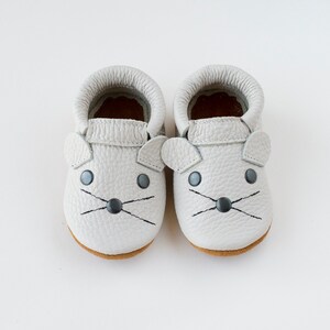 Peut inclure: Une paire de mocassins pour b&eacute;b&eacute; en cuir gris avec un motif de visage de souris. Les mocassins ont des yeux noirs, un nez noir et des moustaches noires.