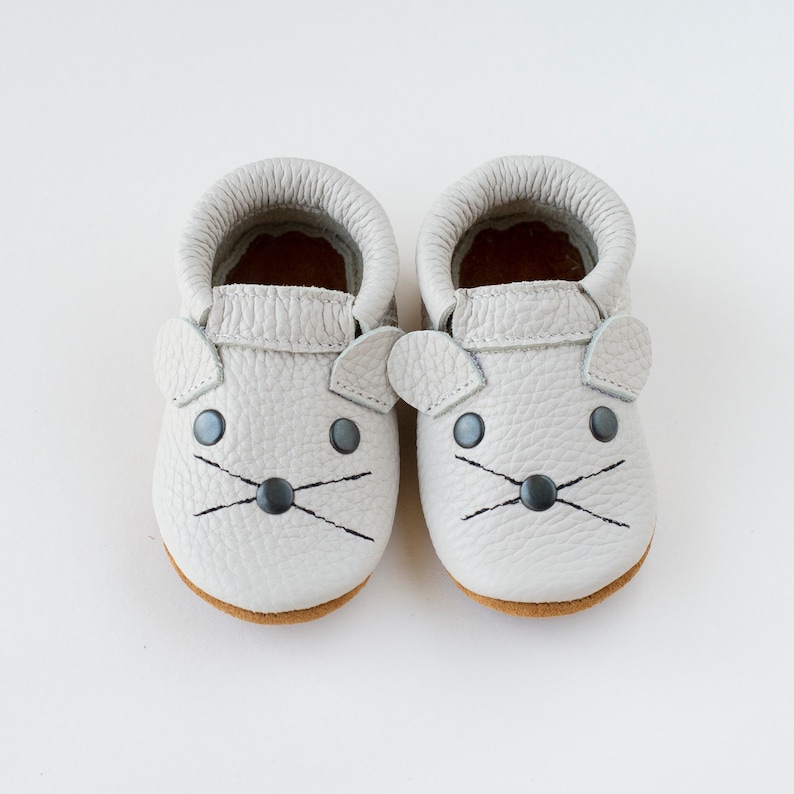Peut inclure: Une paire de mocassins pour b&eacute;b&eacute; en cuir gris avec un motif de visage de souris. Les mocassins ont des yeux noirs, un nez noir et des moustaches noires.