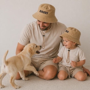 May include: Tan bucket hats with black text reading "DADDY" and "BO" are worn by a man and a child. They are sitting on the floor with a tan puppy. The hats have adjustable chin straps. The man and child are wearing matching tan outfits.