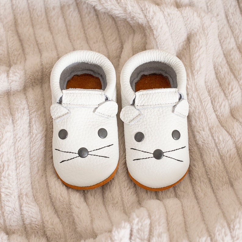 Peut inclure: Une paire de mocassins pour b&eacute;b&eacute; en cuir blanc avec un joli motif de visage de souris. Les mocassins ont des coutures noires pour les yeux, le nez et les moustaches.