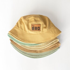 May include: A stack of four bucket hats in various shades of green and yellow. The top hat has a brown leather patch with the text "big bro" in black.