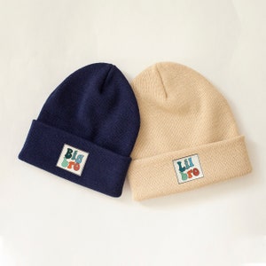 Peut inclure: Deux bonnets en tricot, un bleu marine et un beige, avec du texte coloré. Le bonnet bleu marine dit "Big Bro" et le bonnet beige dit "Lil Bro".