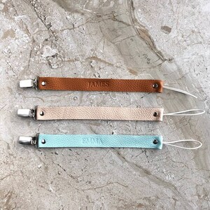 Puede incluir: Tres clips para chupete de cuero con nombres personalizados. Los clips son marrones, beige y azul claro. Cada clip tiene una cuerda blanca.