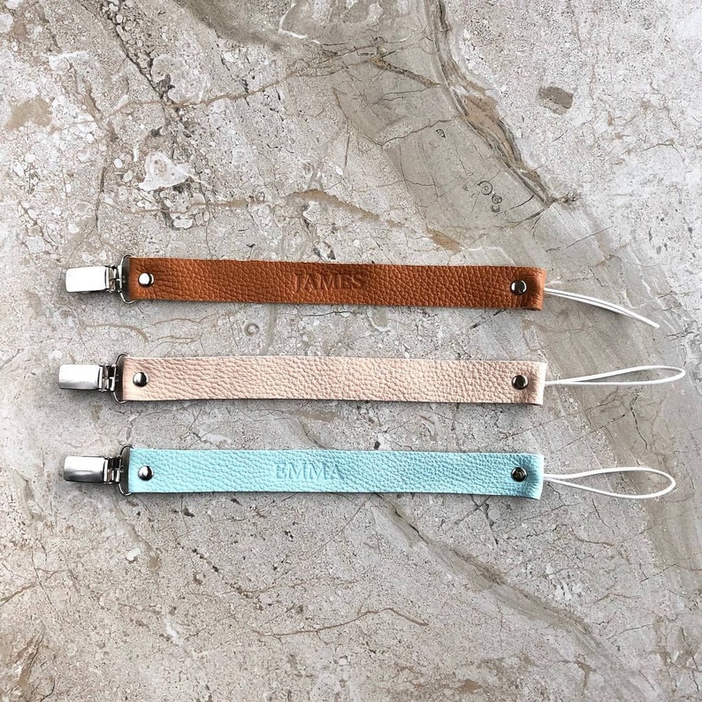Puede incluir: Tres clips para chupete de cuero con nombres personalizados. Los clips son marrones, beige y azul claro. Cada clip tiene una cuerda blanca.