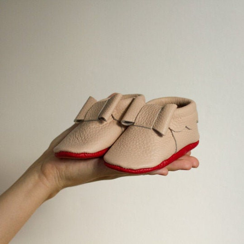 Pu&ograve; includere: Un paio di scarpine per neonati in pelle marrone chiaro con rifiniture rosse. Le scarpe hanno un dettaglio a fiocco sulla parte superiore.