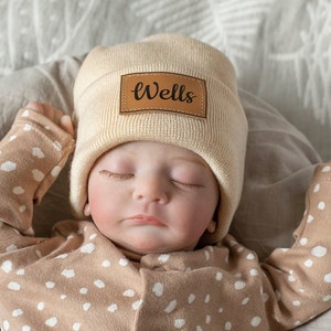 Puede incluir: Un bebé con un gorro de punto beige con un parche de cuero marrón que dice "Wells". El bebé lleva un mono beige con lunares blancos.