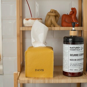 May include: A yellow leather tissue box cover with the word "DAVIS" embossed on the front. The tissue box is on a wooden shelf with a bottle of body butter and decorative items.