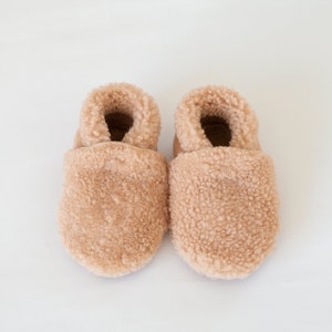 May include: A pair of light brown baby booties with a fluffy, textured exterior. The interior of the booties is a darker brown. The booties are made of a soft, plush material, and are designed to keep little feet warm.
