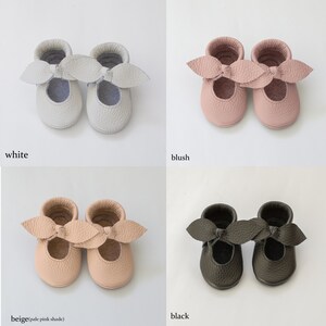 May include: Four pairs of leather baby shoes in different colours: white, blush, beige, and black. Each shoe has a bow on the top.