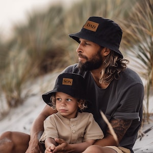 May include: A father and son wearing black bucket hats with brown leather patches that say "DAD" and "SON". The father has a beard and is wearing a black t-shirt. The son is wearing a beige t-shirt.