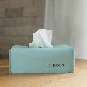 Puede incluir: Una funda para caja de pañuelos de color azul claro con el nombre "JOHNSON" impreso en la parte delantera. La funda está hecha de un material texturizado y tiene una ranura para pañuelos.