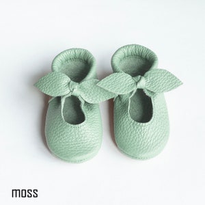 May include: A pair of light green leather baby shoes with a bow on the front. The shoes are called "moss".