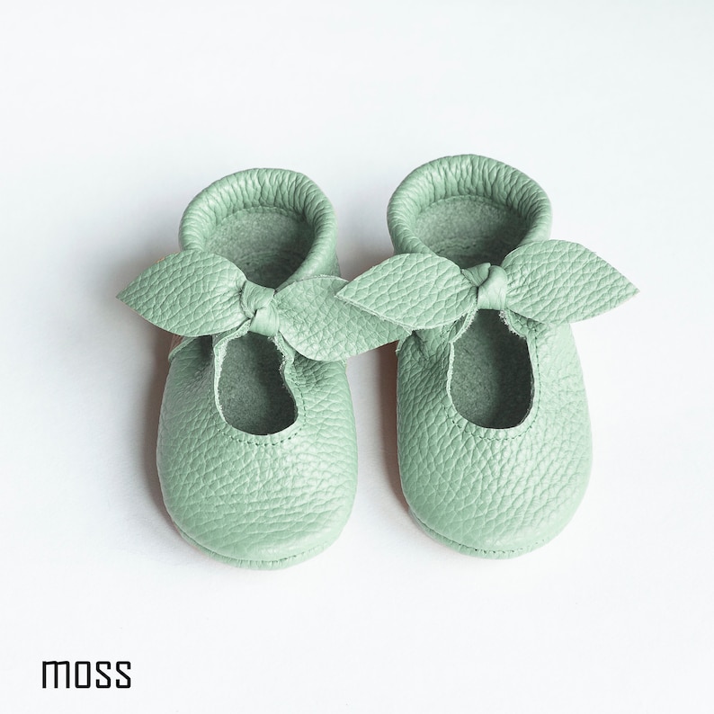 May include: A pair of light green leather baby shoes with a bow on the front. The shoes are called "moss".
