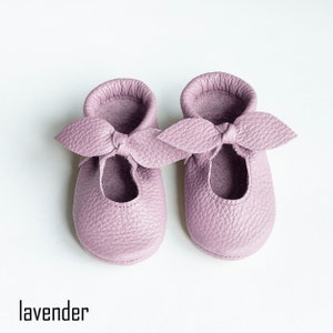 May include: A pair of lavender leather baby shoes with a bow on the front.