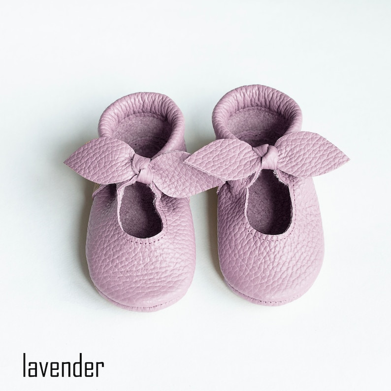 May include: A pair of lavender leather baby shoes with a bow on the front.