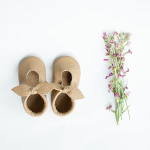 May include: A pair of light brown leather baby shoes with a bow detail. The shoes are on a white background.