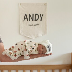 May include: A white baby hat with the name "Andy" embroidered on it. The hat is on a baby wearing a white and brown onesie with a woodland animal print.