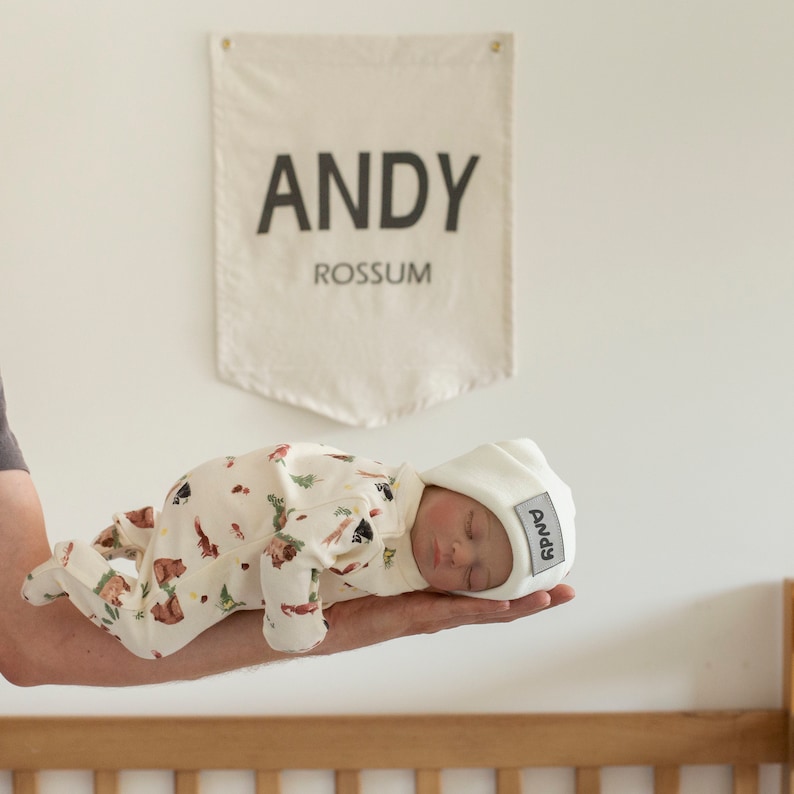 May include: A white baby hat with the name "Andy" embroidered on it. The hat is on a baby wearing a white and brown onesie with a woodland animal print.